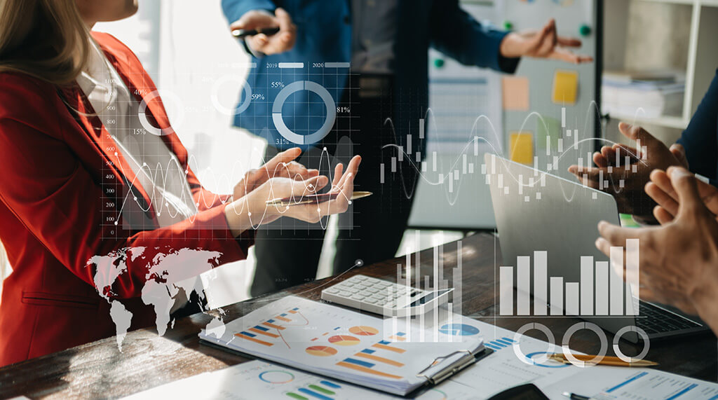 Três profissionais de frente para seus respectivos notebooks. Entre os notebooks, há diversos papéis com gráficos. No canto esquerdo para o centro da imagem, há gráficos virtuais com dados corporativos.