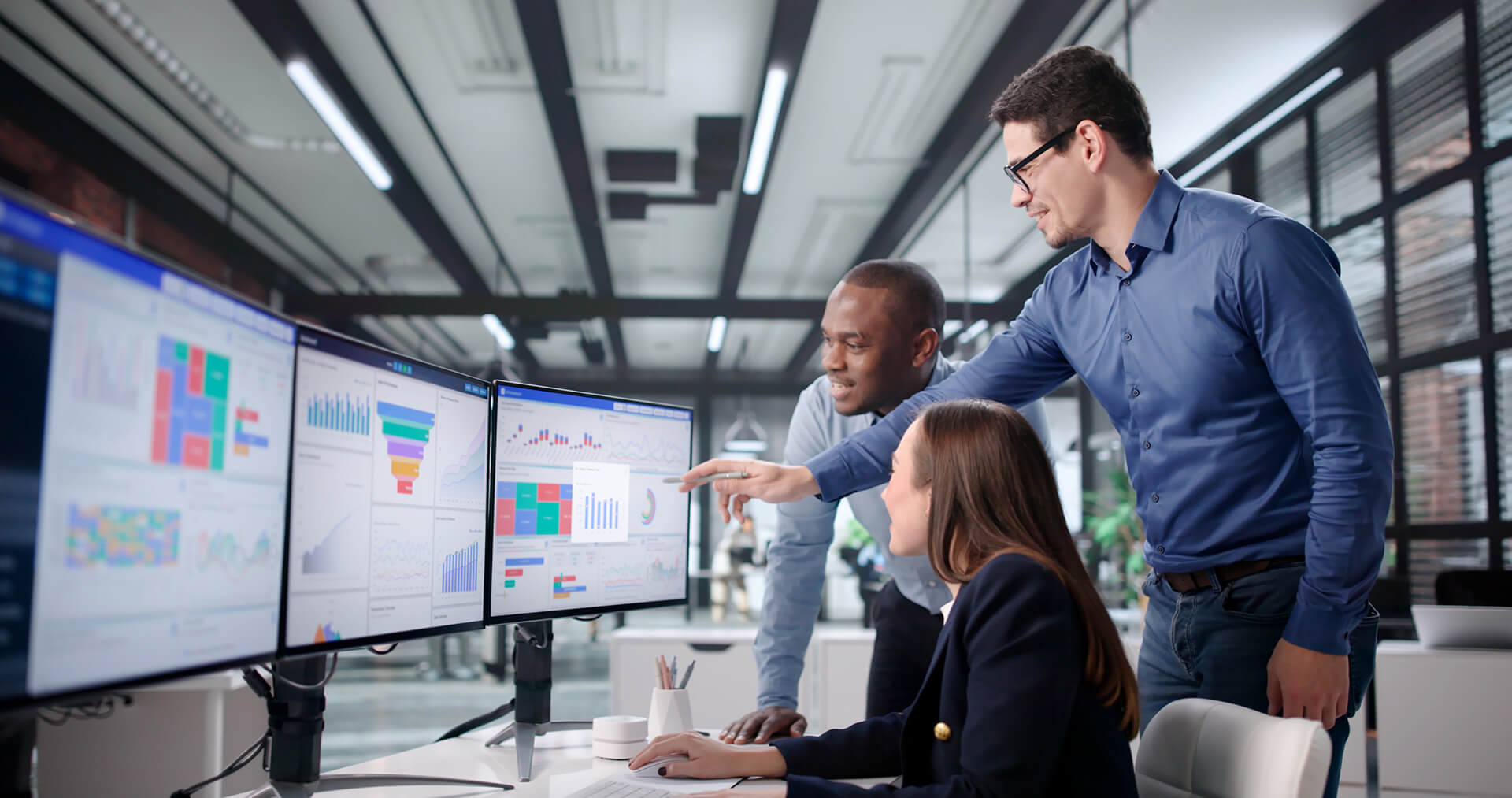 Três profissionais (dois homens e uma mulher) em frente a três monitores com gráficos de KPI.